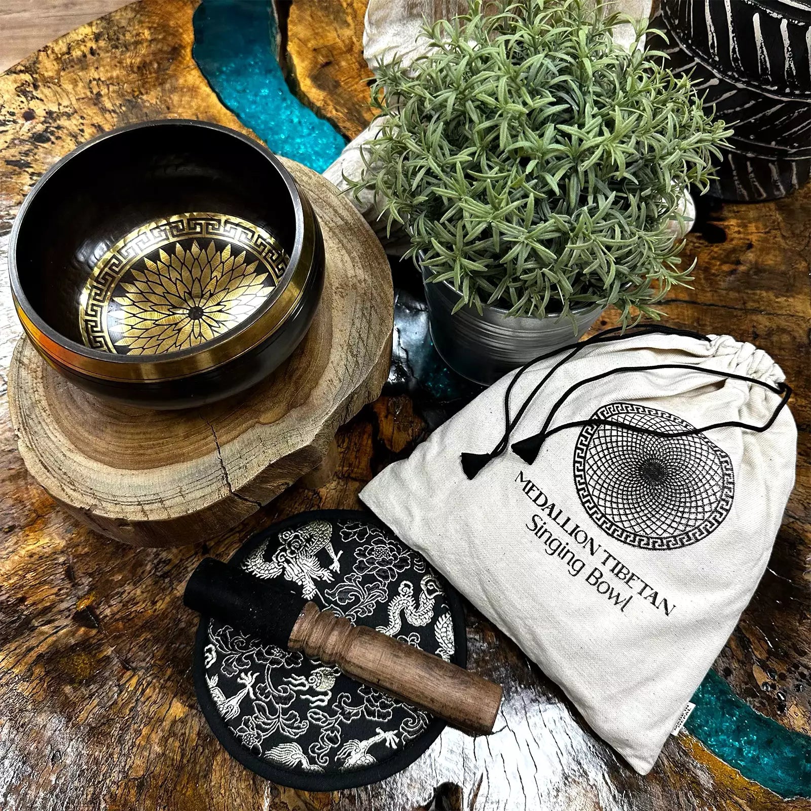 A singing bowl set arranged on a rustic wooden surface, including a black metal singing bowl with a gold-toned engraved interior, a white drawstring pouch printed with 'MEDULLION THERAPY Singing Bowl,' a black-and-white patterned cloth, and a wooden-handled striker. A potted green plant and decorative stones are also present.