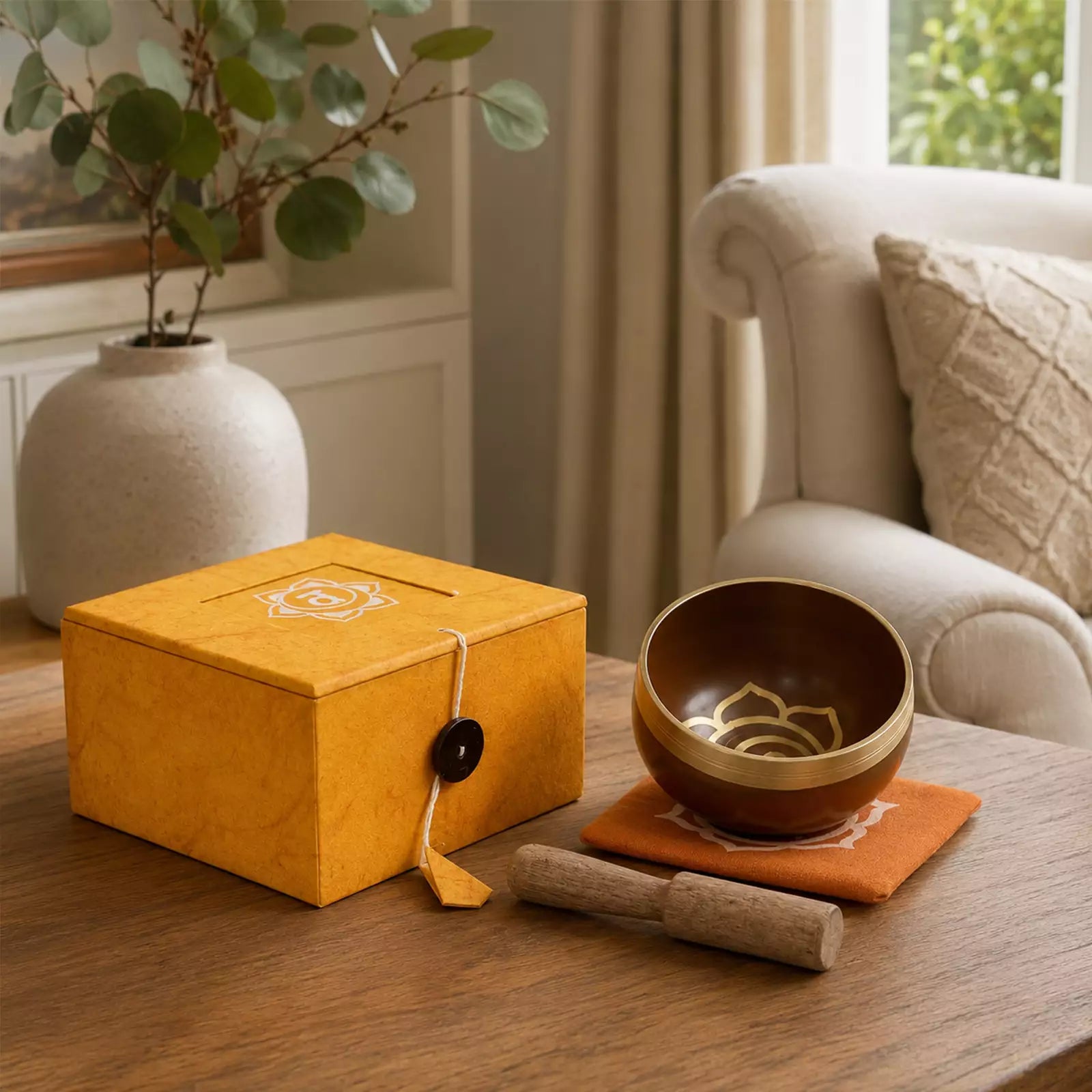 A wooden singing bowl set with a metallic striker and a decorative orange cloth, styled on a wooden table next to a yellow gift box in a cozy living room setting.