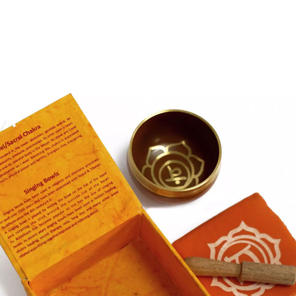 A brass singing bowl set with an orange box, a folded orange cloth, and a wooden striker on a white background. The inside of the box shows printed instructions and the word 'Chakra'.