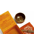 A brass singing bowl set with an orange box, a folded orange cloth, and a wooden striker on a white background. The inside of the box shows printed instructions and the word 'Chakra'.