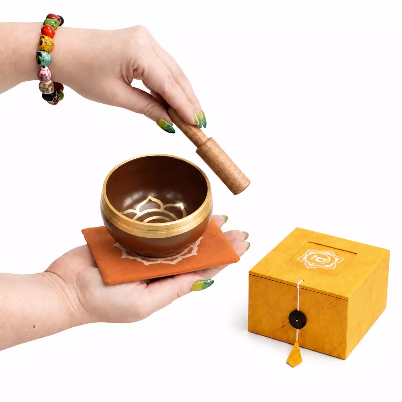 A person holds a small singing bowl with a wooden striker above an orange coaster, with a yellow gift box beside it on a white background.