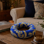 A blue and gold patterned circular cushion or ring-shaped pillow sits on a wooden table in a cozy living room setting with a sofa, books, and candles in the background.