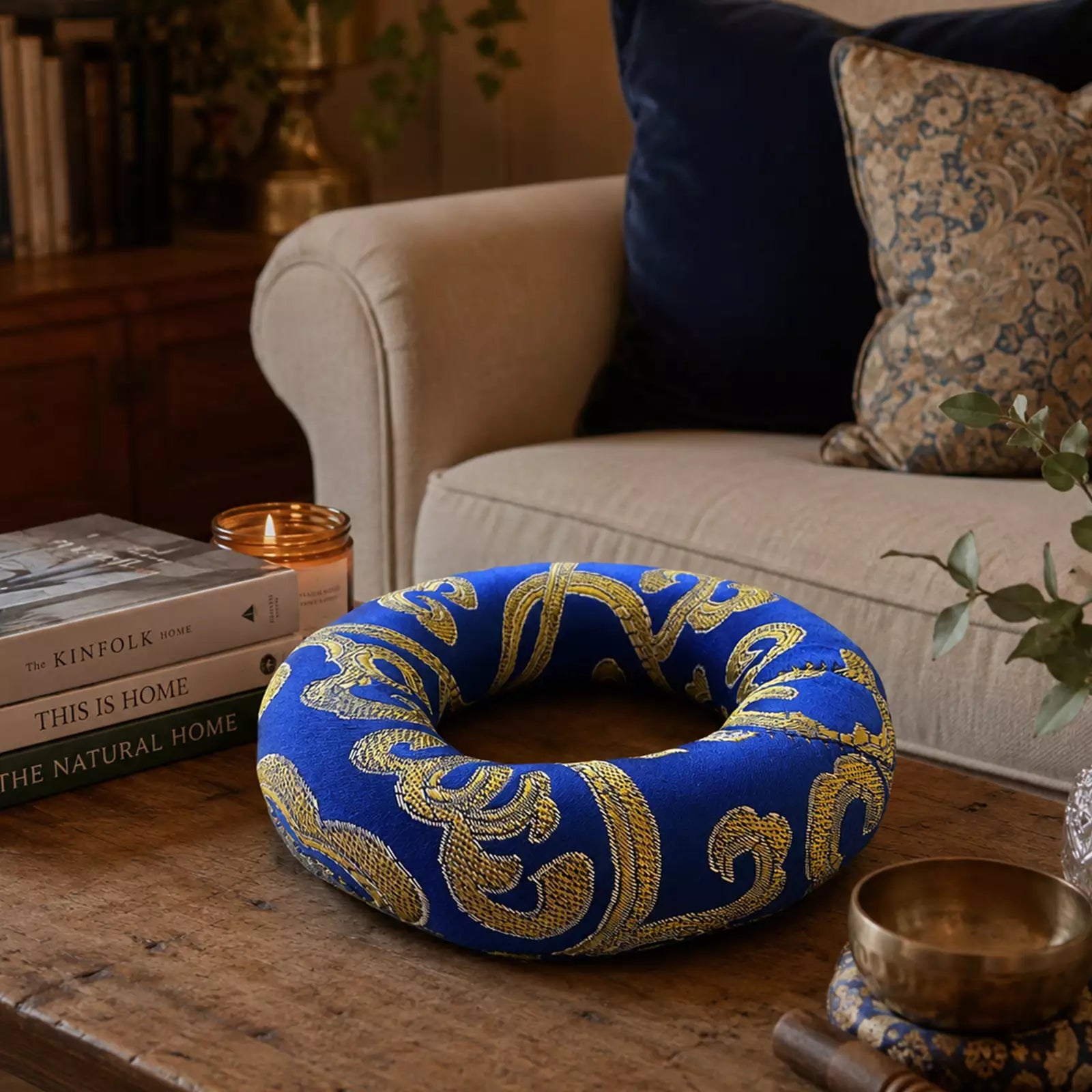 A blue and gold patterned circular cushion or ring-shaped pillow sits on a wooden table in a cozy living room setting with a sofa, books, and candles in the background.