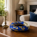 A blue and gold patterned donut-shaped decorative object sits on a wooden table in a styled living room setting with a sofa, lamp, and fireplace visible in the background.