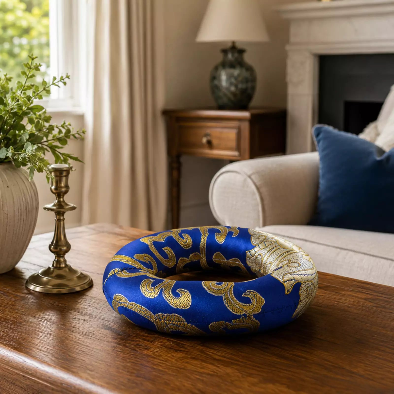 A blue and gold patterned donut-shaped decorative object sits on a wooden table in a styled living room setting with a sofa, lamp, and fireplace visible in the background.