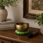 A brass singing bowl with a green and gold patterned cushion and a wooden mallet resting on a wooden table in a styled home interior setting.