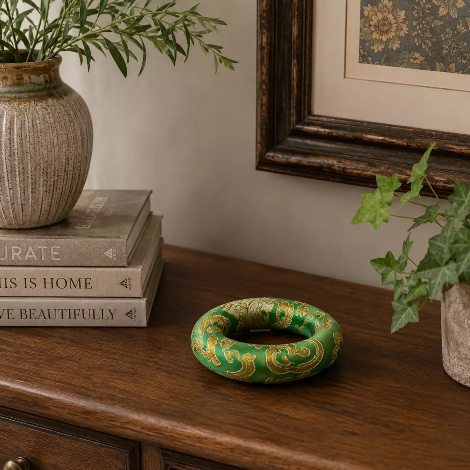 A green and gold patterned ring-shaped decorative object sits on a wooden console table in a styled interior setting with books, a vase, framed wall art, and ivy.