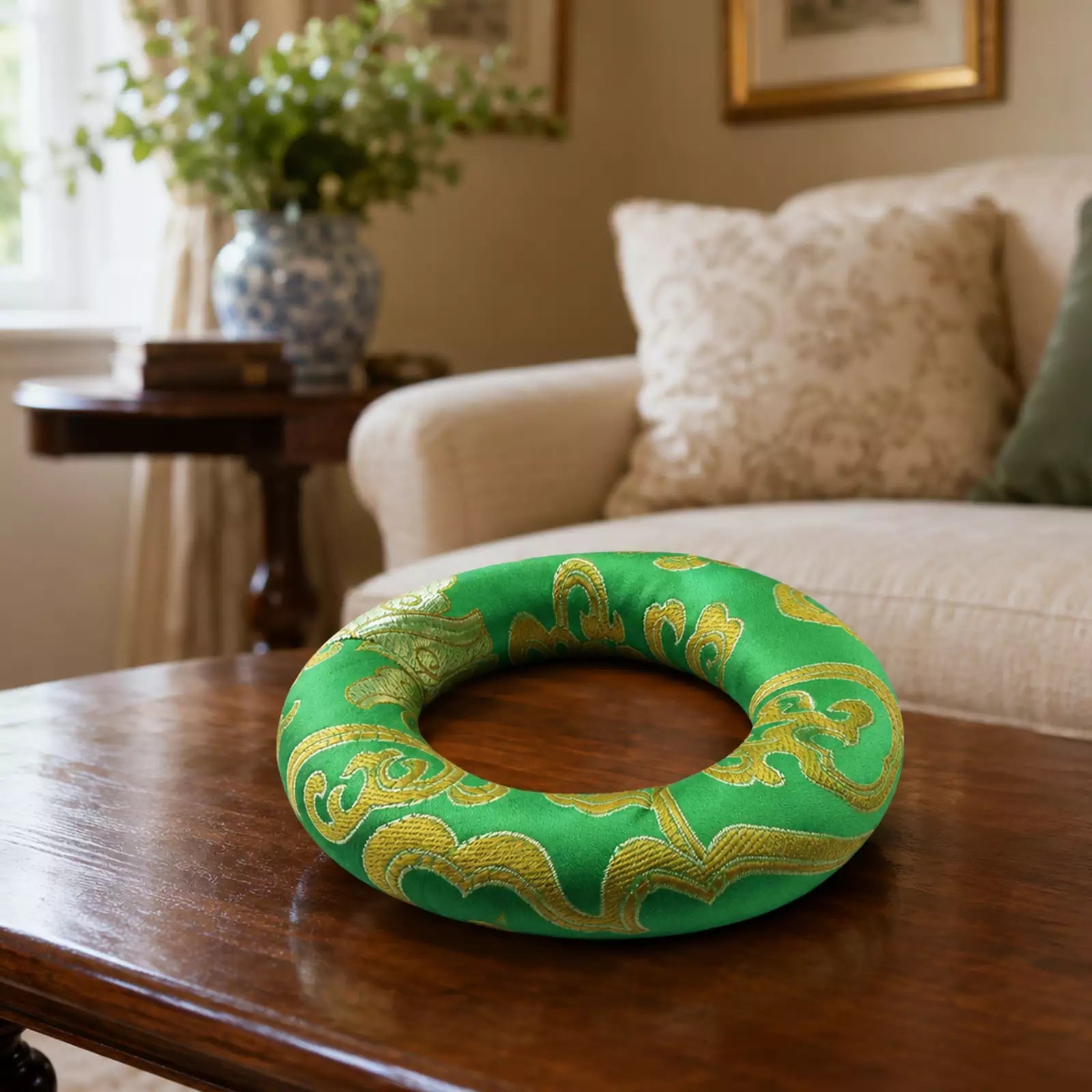 A green fabric ring cushion with gold embroidered wave patterns, resting on a dark wooden coffee table in a cozy living room setting.