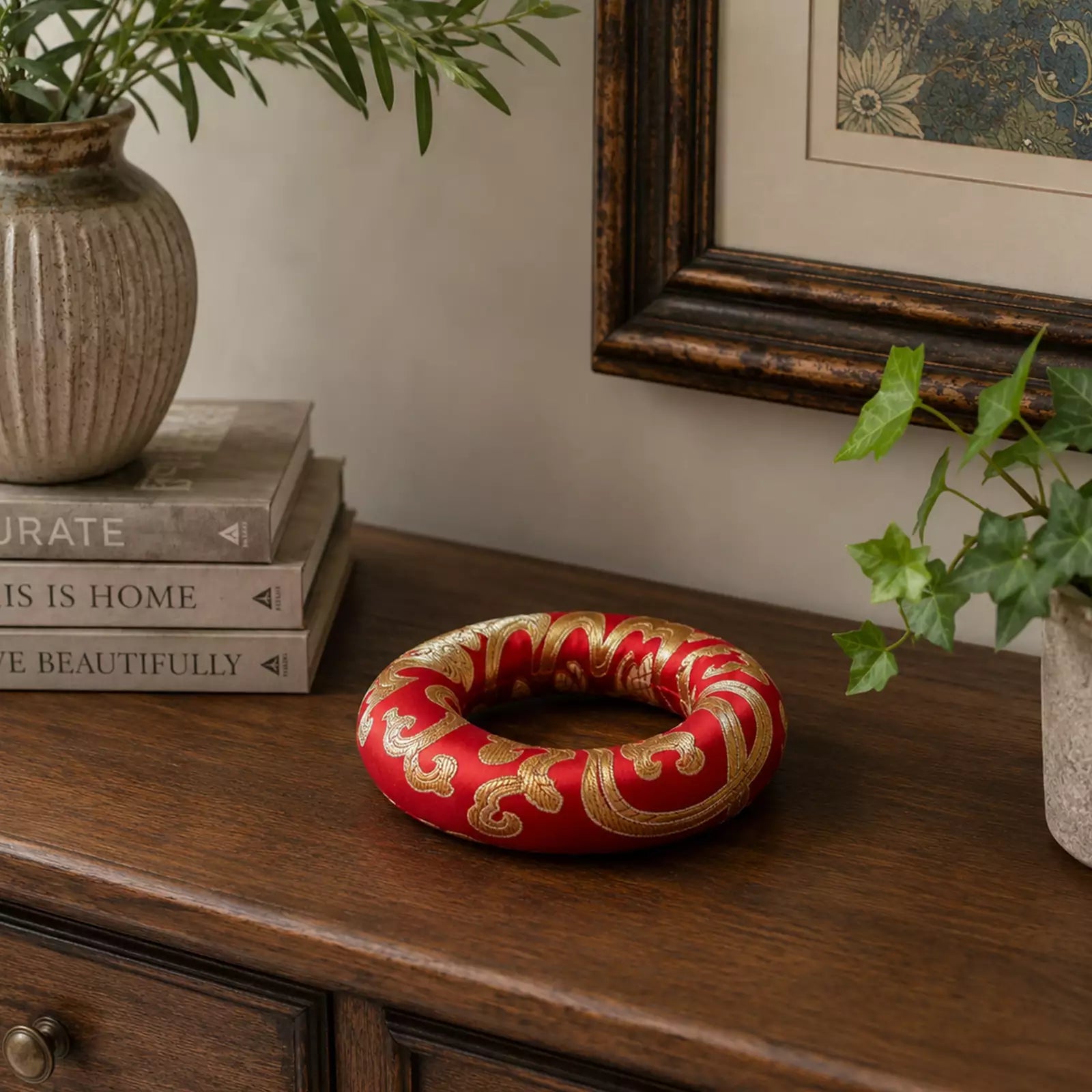 A red fabric ring-shaped decorative piece with ornate gold embroidery sits on a dark wooden console table in a styled room setting with books, potted plants, and a framed picture.