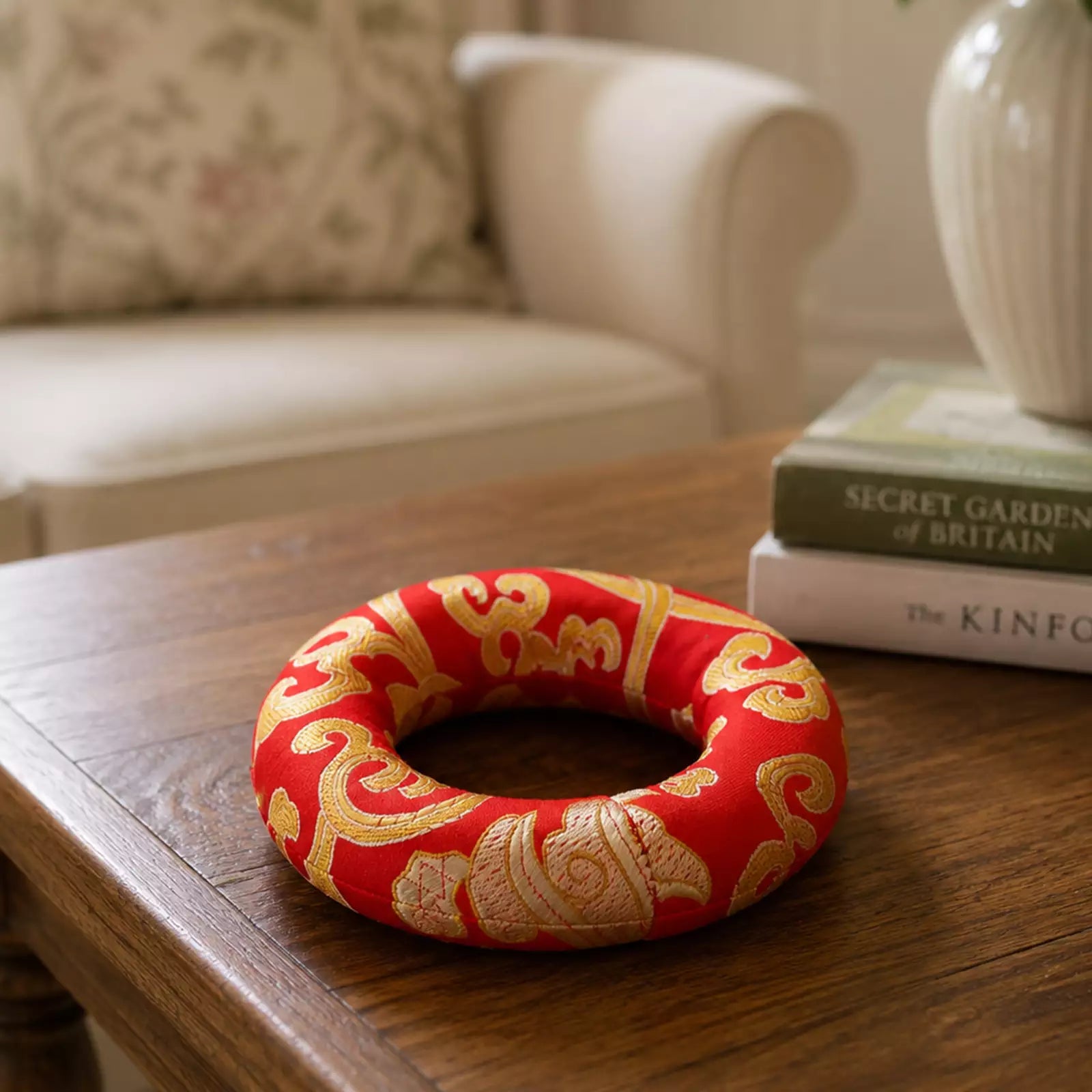 A red fabric ring with gold ornamental patterns sits on a wooden table in a softly lit indoor setting, with books and a chair blurred in the background.