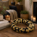 A black and gold patterned decorative wreath displayed on a wooden table in a cozy living room setting with candles, books, and plants.