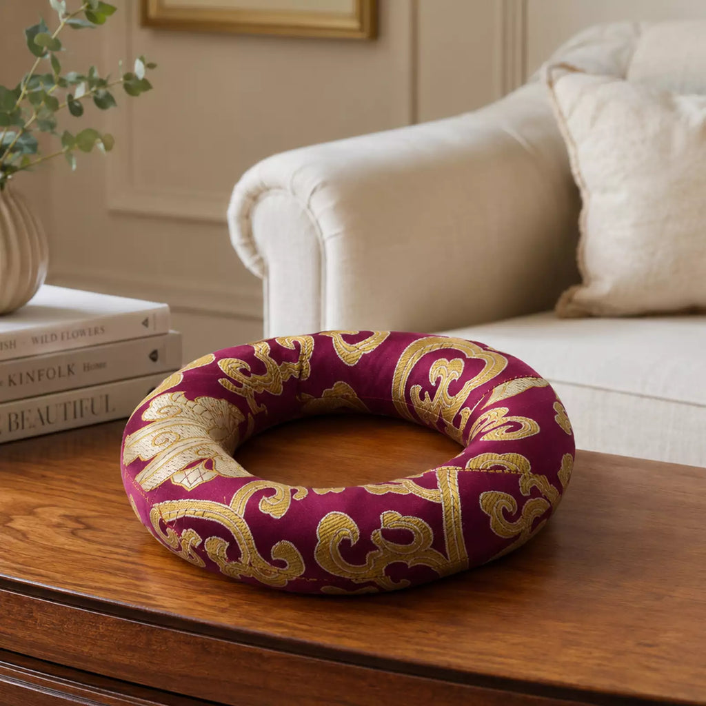 A circular decorative wreath with a deep burgundy fabric and ornate gold scrollwork, displayed on a wooden table in a living room setting.