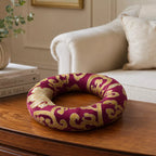 A circular decorative wreath with a deep burgundy fabric and ornate gold scrollwork, displayed on a wooden table in a living room setting.