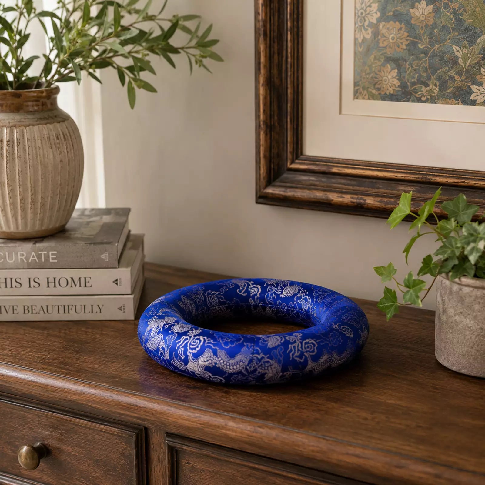 A blue and white patterned ceramic ring-shaped planter sits on a dark wooden console table in a styled room setting with books, a potted plant, and framed wall art.