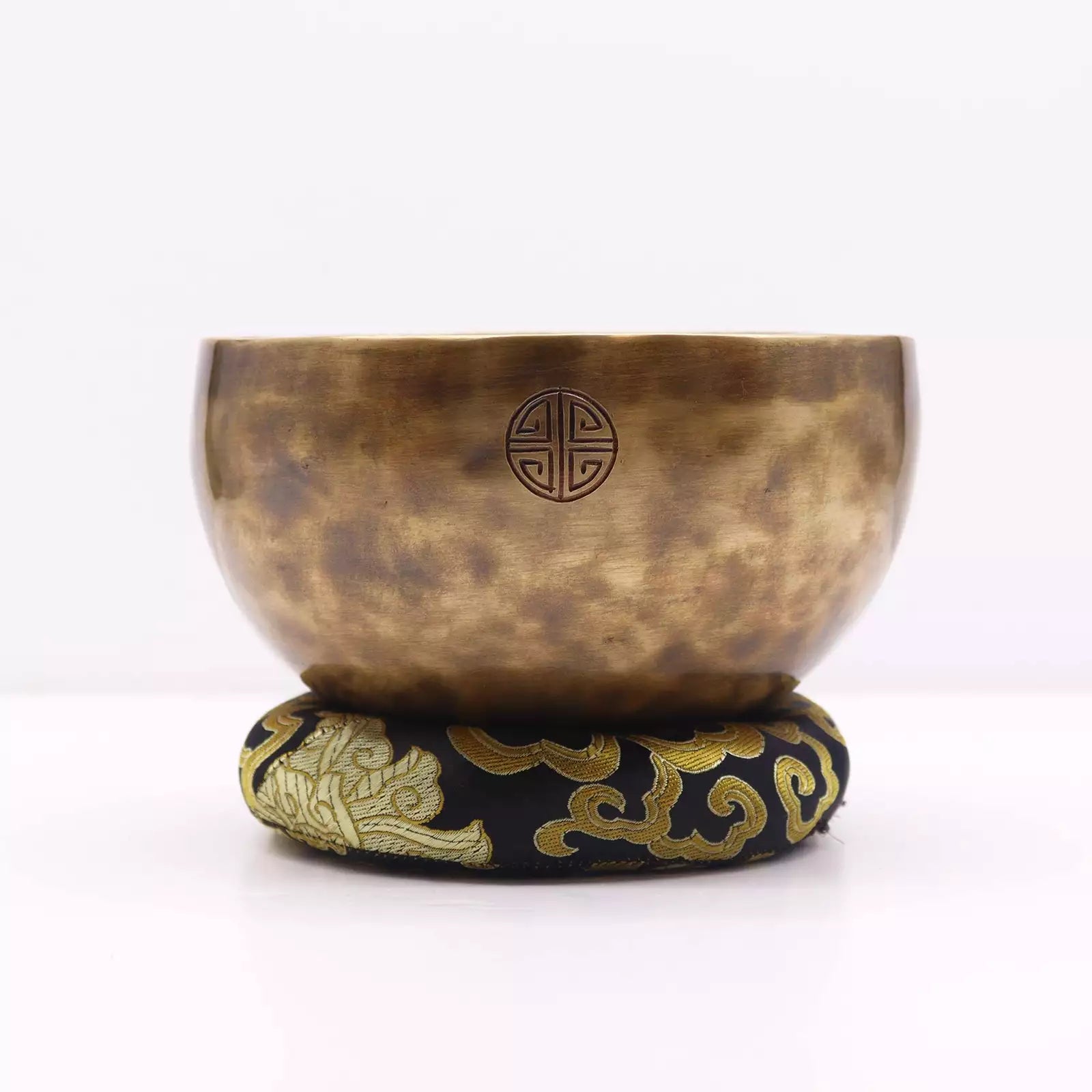 A bronze singing bowl with a hammered texture and a Chinese geometric emblem, resting on a black and gold embroidered cushion against a white background.