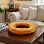 An orange and gold patterned circular cushion or ring-shaped pillow displayed on a dark wooden coffee table in a living room setting.