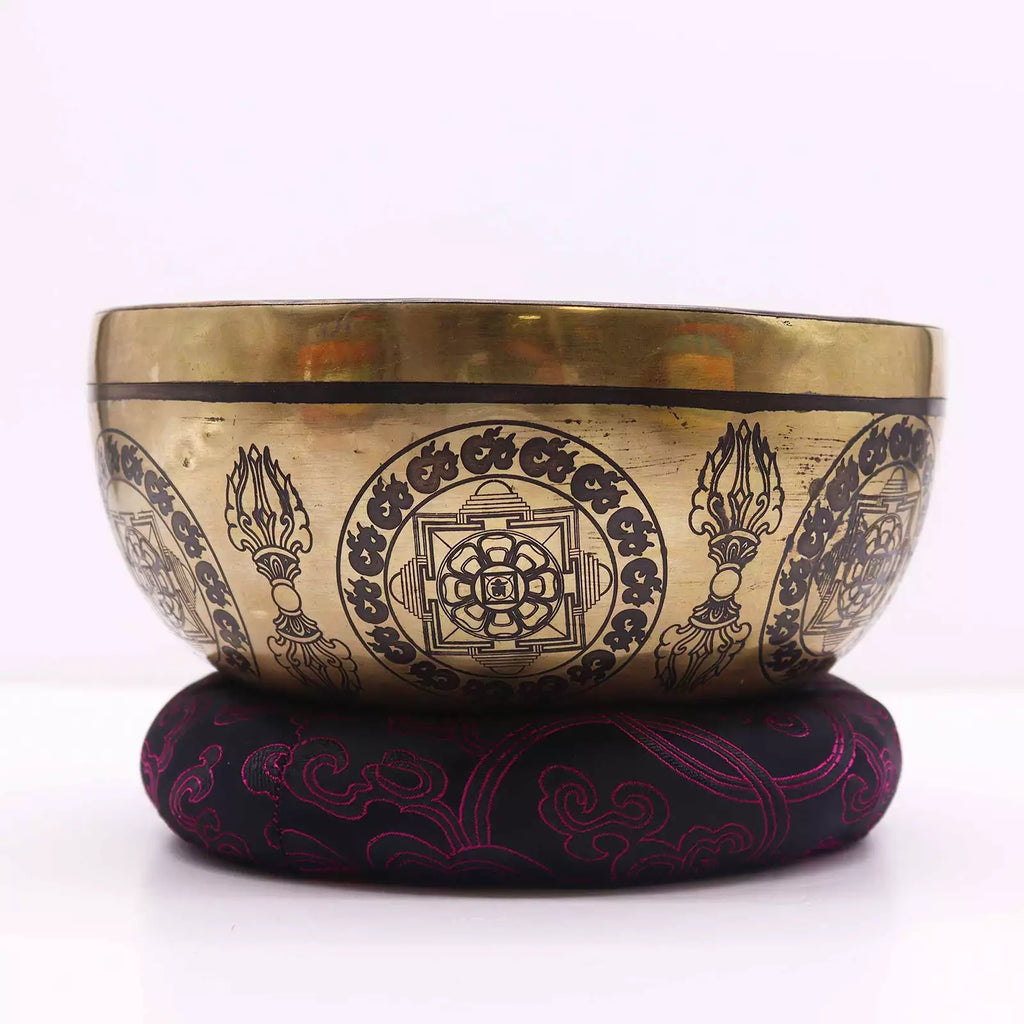 A Tibetan singing bowl made of gold-toned metal with black engraved patterns, resting on a round black cushion with magenta embroidery, set against a plain white background.