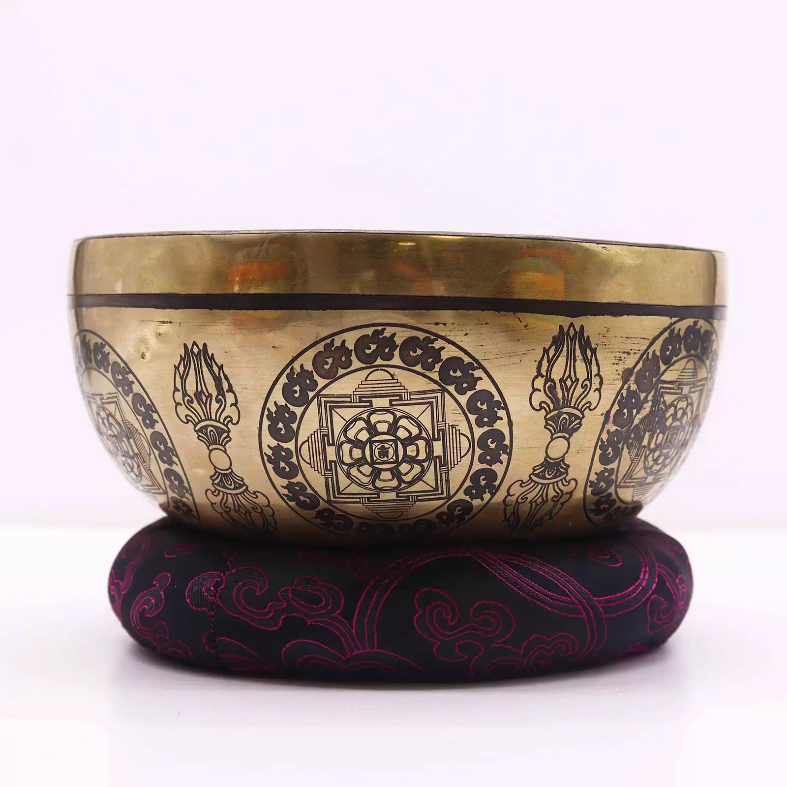 A Tibetan singing bowl made of gold-toned metal with black engraved patterns, resting on a round black cushion with magenta embroidery, set against a plain white background.