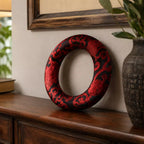 A decorative circular ring with a red and black damask pattern, displayed on a dark wood console table against a beige wall.
