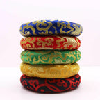 A stack of five round cushion covers or small ottomans made of ornate brocade fabric, shown on a plain light background. The colors from top to bottom are blue, red, green, yellow, and black, each with a gold cloud-like pattern.