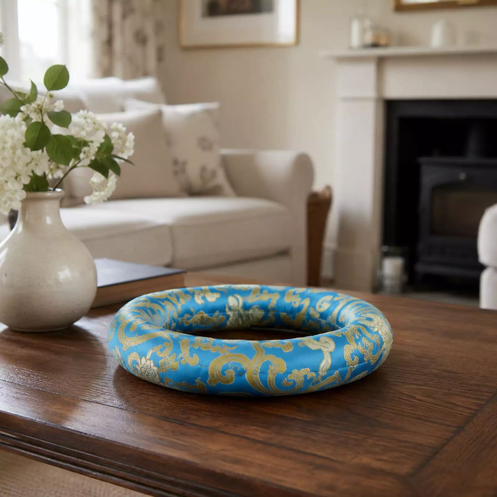 A blue and gold damask-patterned decorative ring or wreath displayed on a dark wooden table in a softly blurred living room setting.
