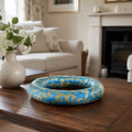 A blue and gold damask-patterned decorative ring or wreath displayed on a dark wooden table in a softly blurred living room setting.