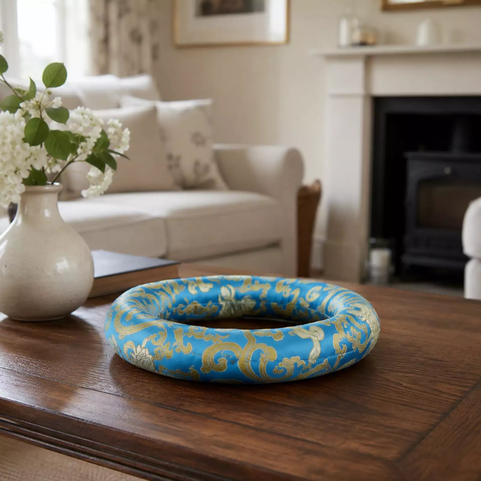 A blue and gold damask-patterned decorative ring or wreath displayed on a dark wooden table in a softly blurred living room setting.