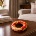 A red and gold decorative ring-shaped ornament with embroidered patterns, resting on a dark wooden table in a softly blurred living room setting.