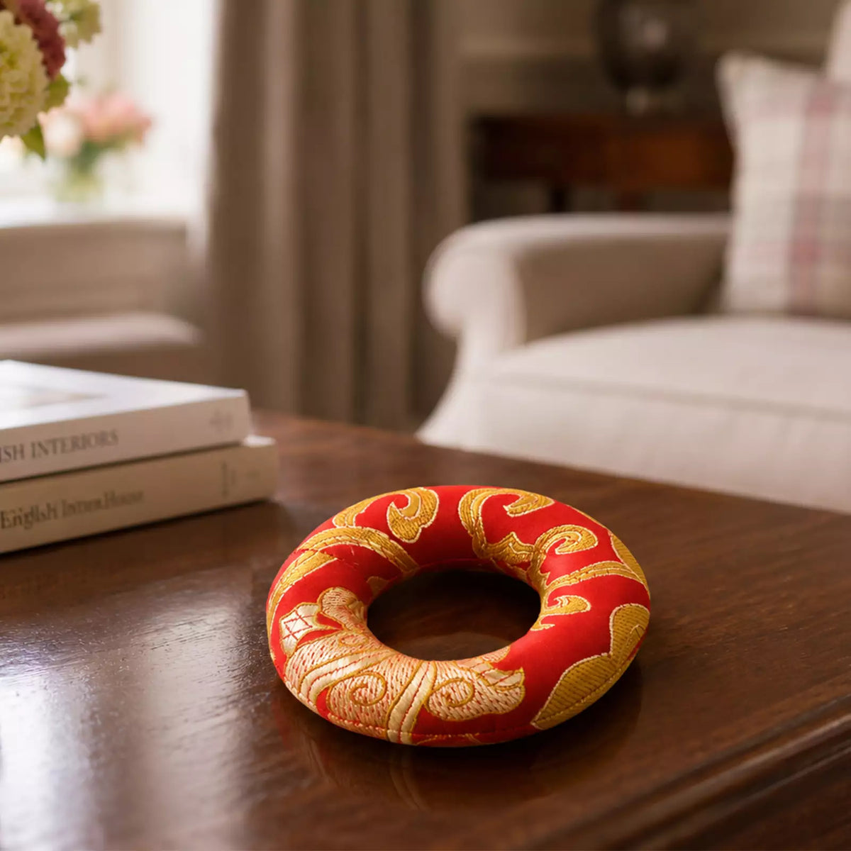 A red and gold decorative ring-shaped ornament with embroidered patterns, resting on a dark wooden table in a softly blurred living room setting.