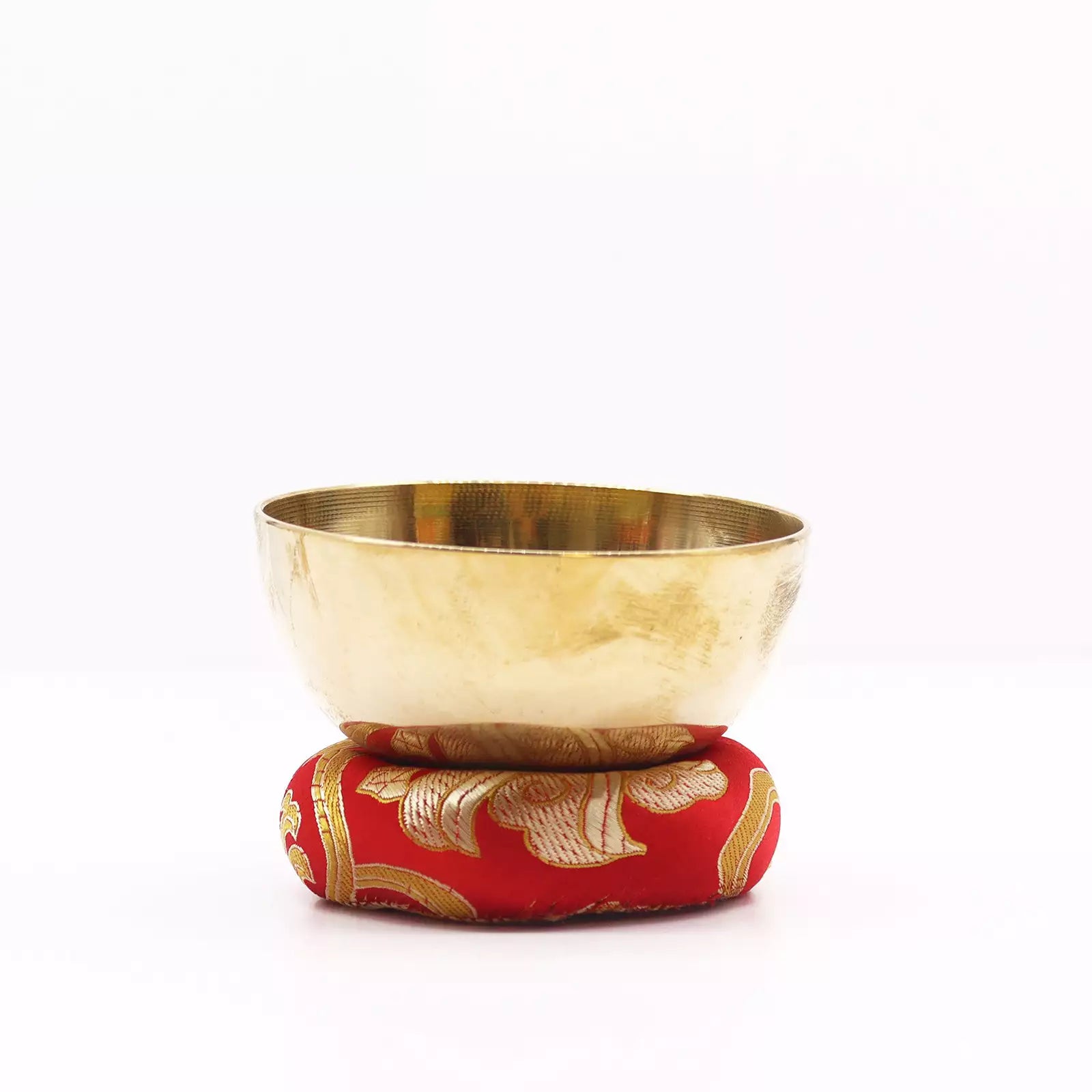 A brass singing bowl resting on a red and gold patterned cushion, photographed against a seamless white background.