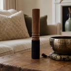 A wooden singing bowl mallet with a dark wrapped handle stands on a rustic wooden table next to a brass singing bowl on a cushion, with a softly blurred living room background.