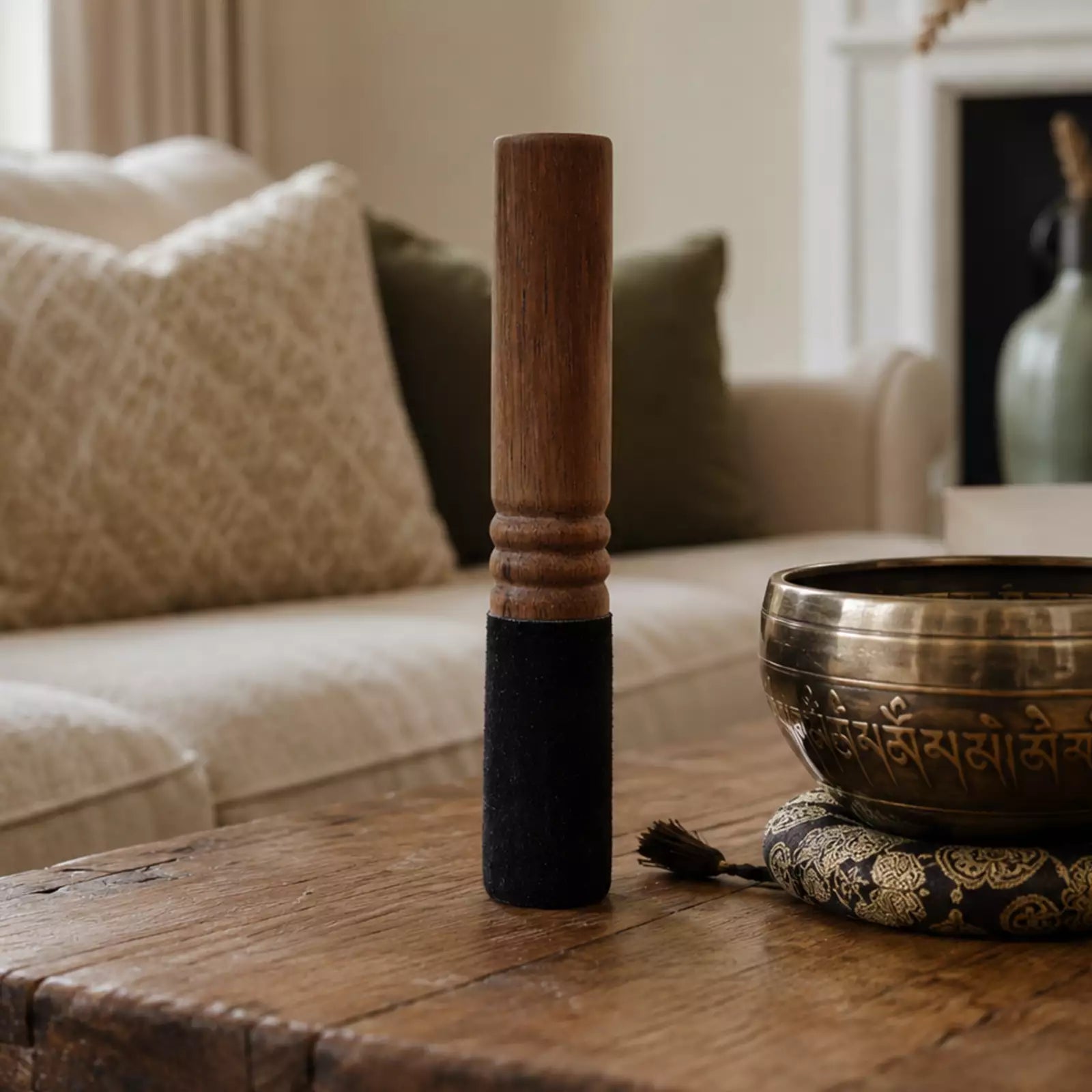 A wooden singing bowl mallet with a dark wrapped handle stands on a rustic wooden table next to a brass singing bowl on a cushion, with a softly blurred living room background.