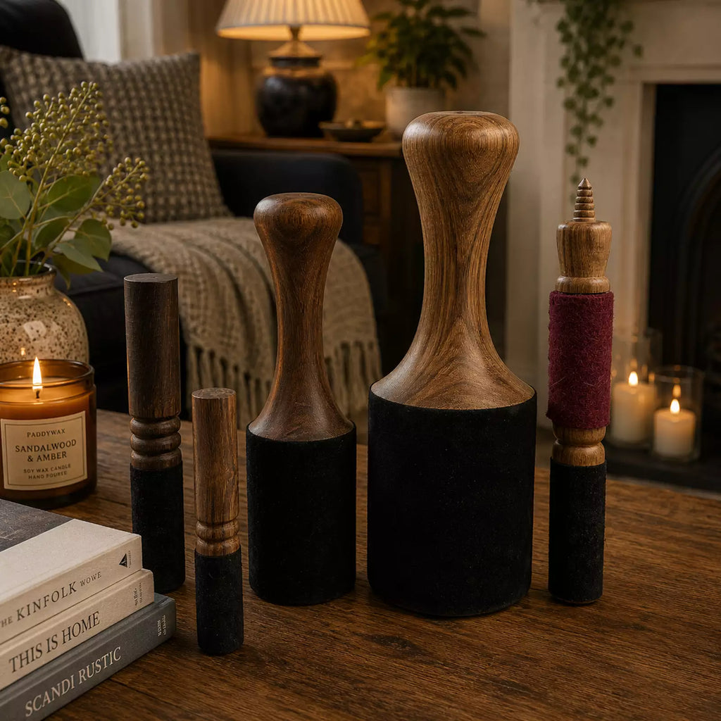 A set of six wooden and fabric massage or rhythmic striking tools arranged on a wooden table in a cozy, warmly lit living room setting with candles, books, and home decor visible in the background.