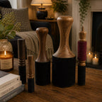 A set of six wooden and fabric massage or rhythmic striking tools arranged on a wooden table in a cozy, warmly lit living room setting with candles, books, and home decor visible in the background.