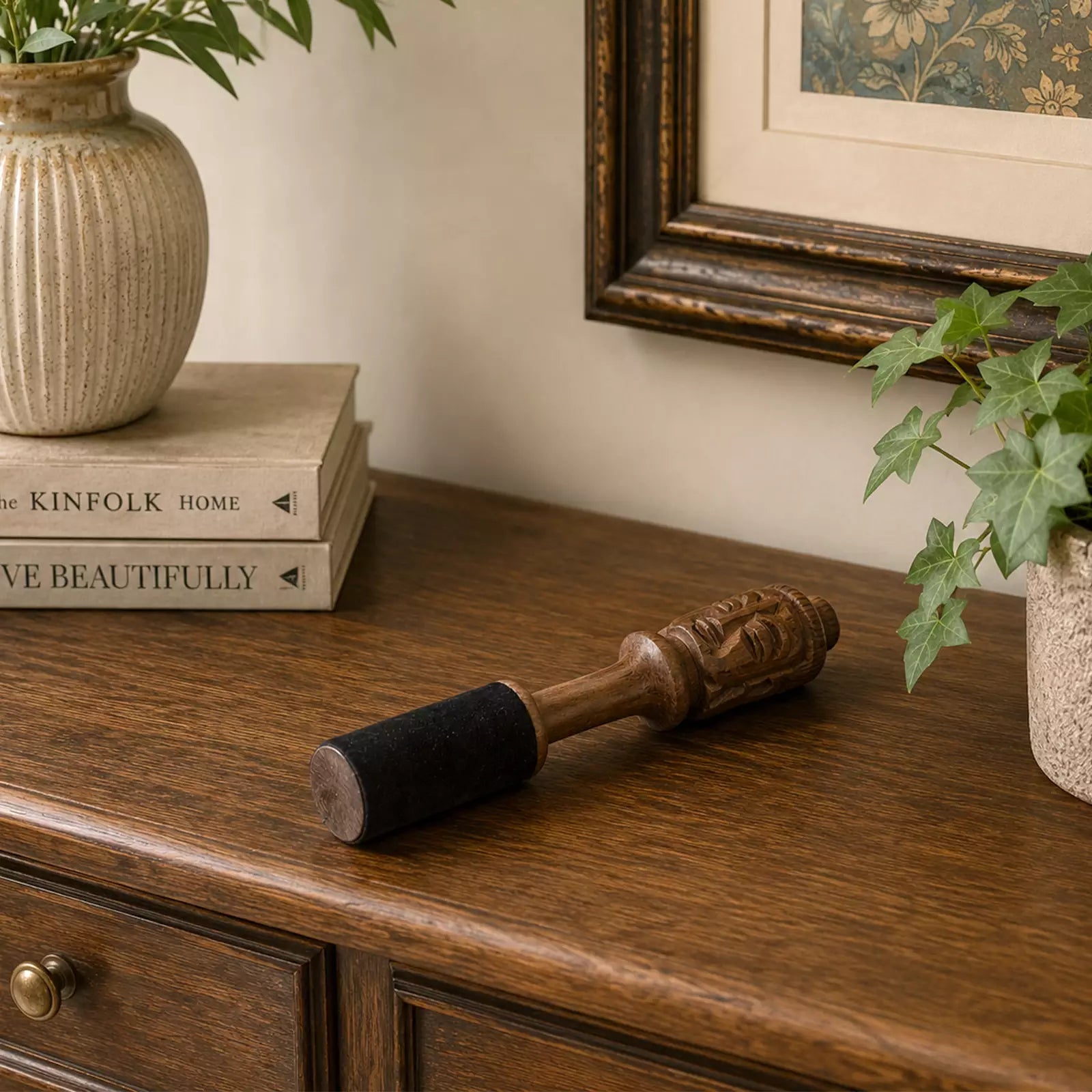 A vintage-style wooden comb rest with a carved handle and black velvet roller, resting on a dark wood sideboard next to books and potted ivy.