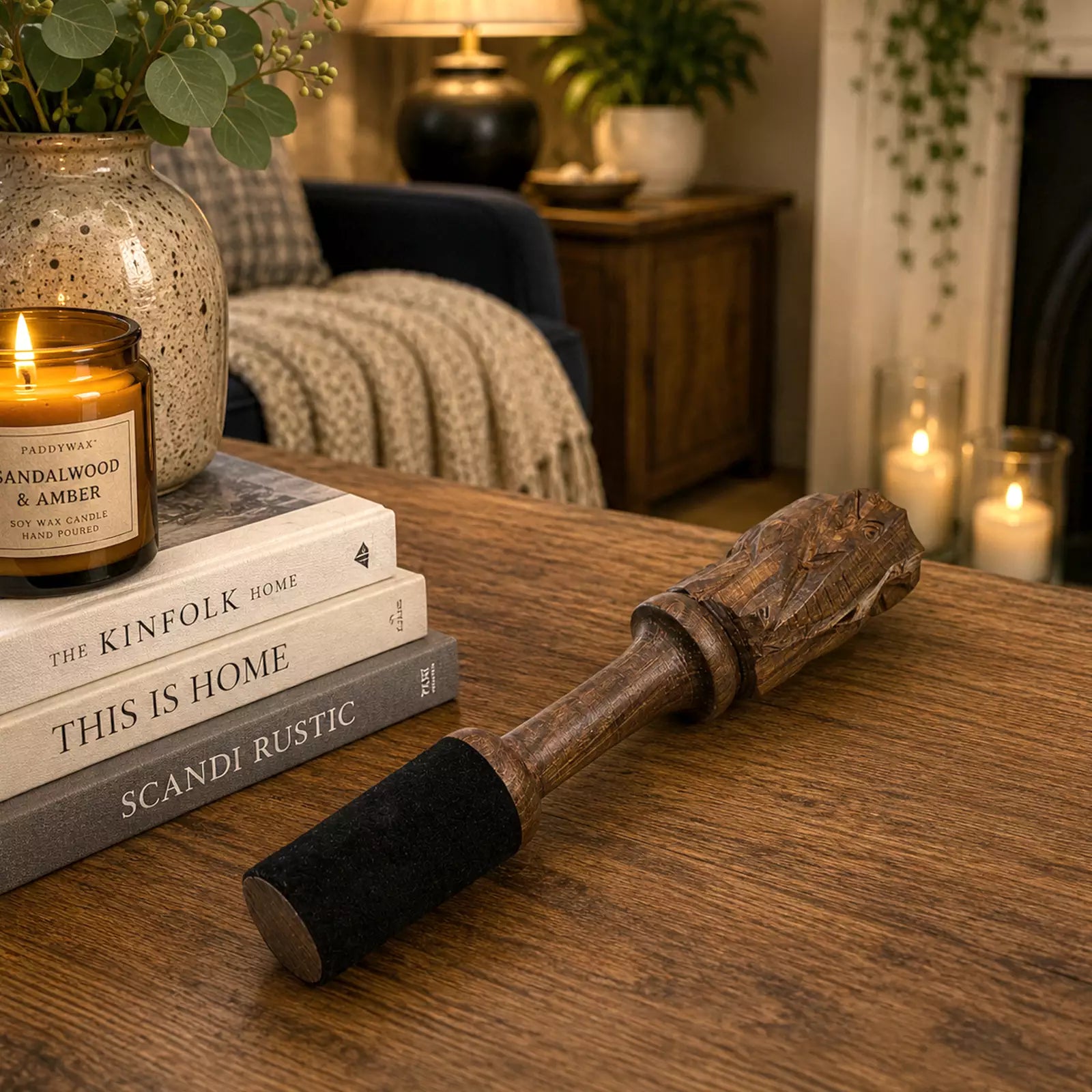 A rustic wooden and black velvet wine bottle opener lies on a wooden table in a cozy, warmly lit living room setting. The background features a bookshelf with a lit candle, a knitted throw blanket, and a fireplace with glowing candles.
