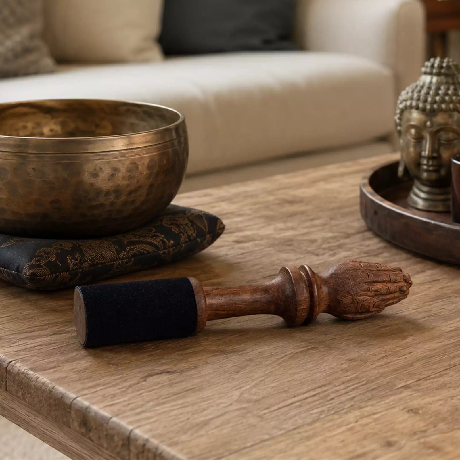 A wooden singing bowl set with a carved wooden striker wrapped in black felt, resting on a rustic wooden table in a cozy living room setting.
