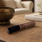 A wooden singing bowl striker with a dark velvet sleeve rests on a wooden table in a cozy living room setting, with a brass singing bowl and a book in the background.