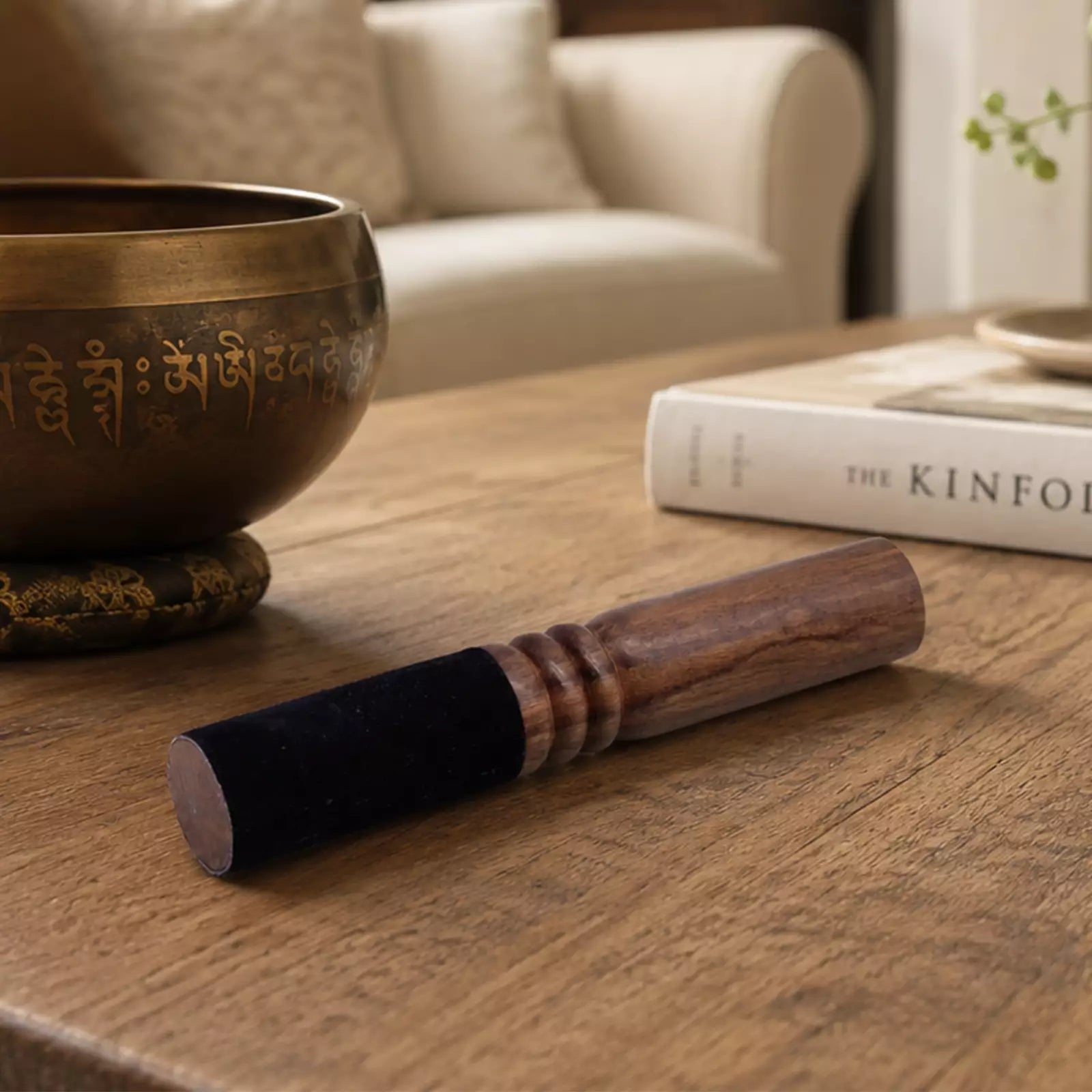 A wooden singing bowl striker with a dark velvet sleeve rests on a wooden table in a cozy living room setting, with a brass singing bowl and a book in the background.