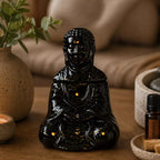 Sitting Buddha Oil Burner - Black in a serene setting with smoke rising from the top, surrounded by calming decor including a candle, plant, and essential oil bottle.