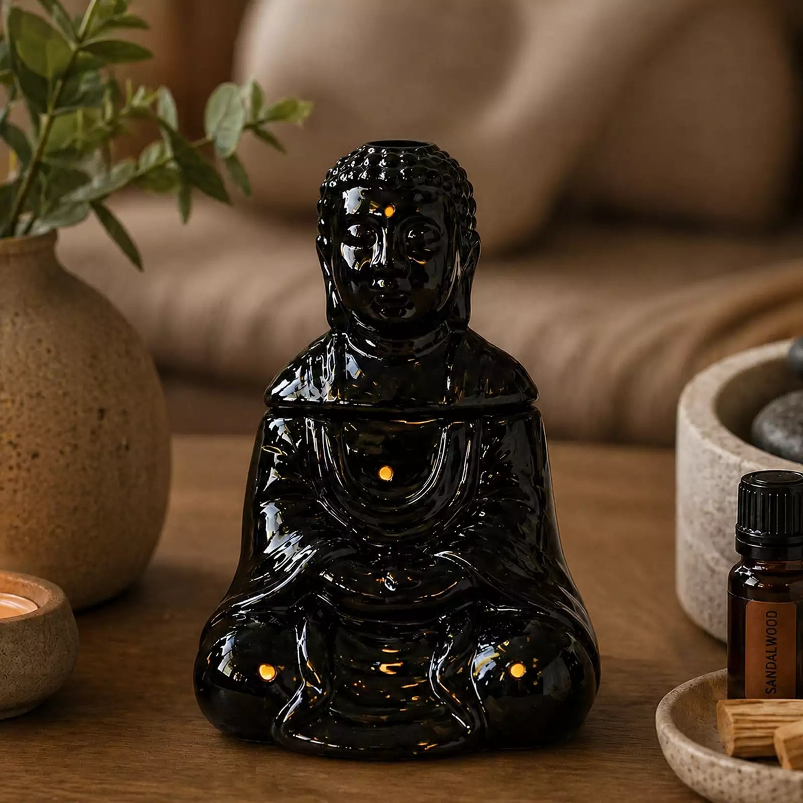 Sitting Buddha Oil Burner - Black in a serene setting with smoke rising from the top, surrounded by calming decor including a candle, plant, and essential oil bottle.
