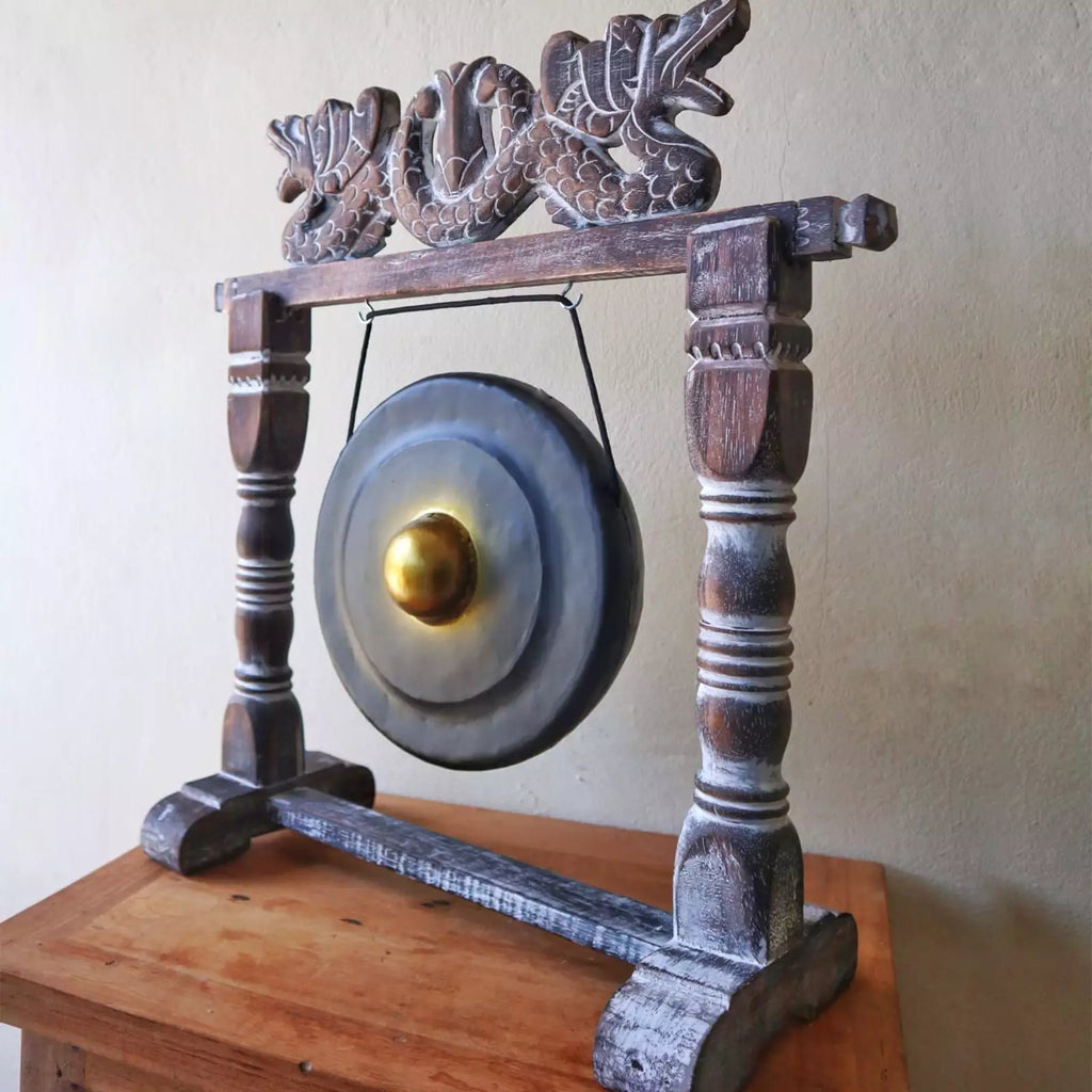 A large round gong with a metallic bronze finish and a polished gold center, mounted on a carved dark wood stand with ornate dragon-like detailing at the top, displayed on a wooden surface against a plain beige wall.