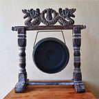 A dark carved wooden gong stand holding a large round black gong, photographed against a plain beige wall with a wooden surface below.