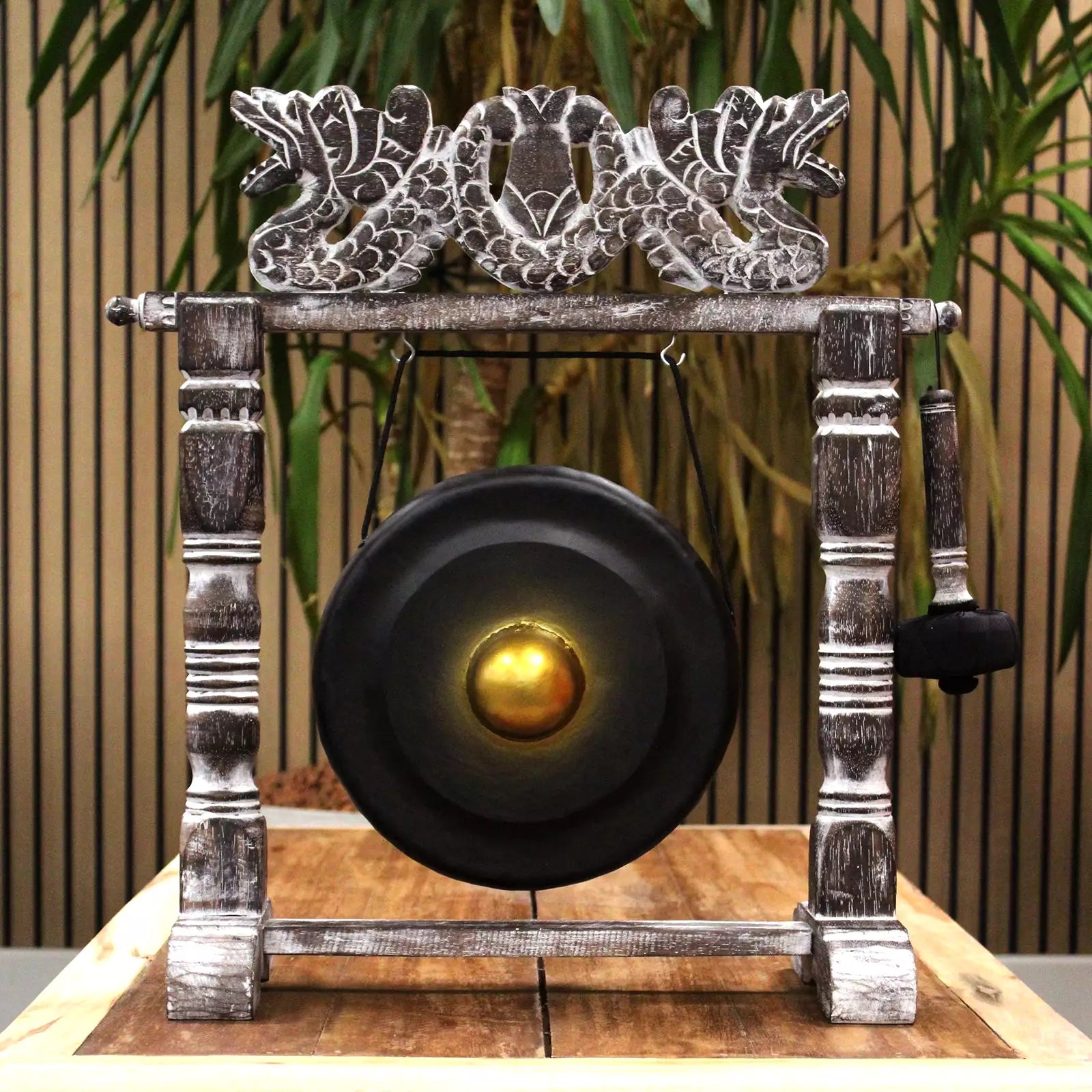A decorative gong set with a large black circular gong, a silver-toned ornate frame, and a hanging mallet, displayed on a wooden surface with a slatted wall and green plants in the background.