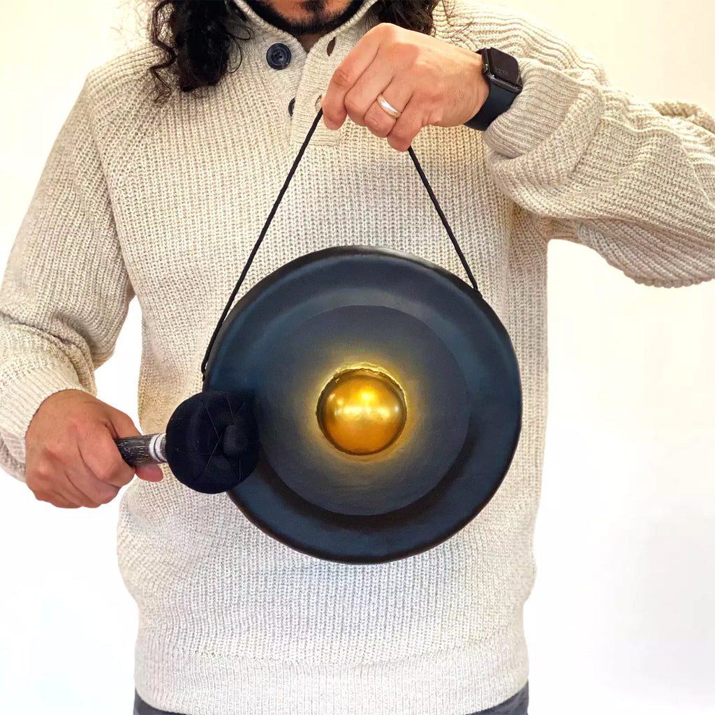 A person wearing a cream knit sweater holds a black hanging gong with a metallic gold center and strikes it with a black mallet against a plain white background.