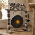 A rustic decorative gong set with a dark round gong, weathered wood stand, carved dragon-like top piece, and a hanging mallet, shown in a warm indoor setting.