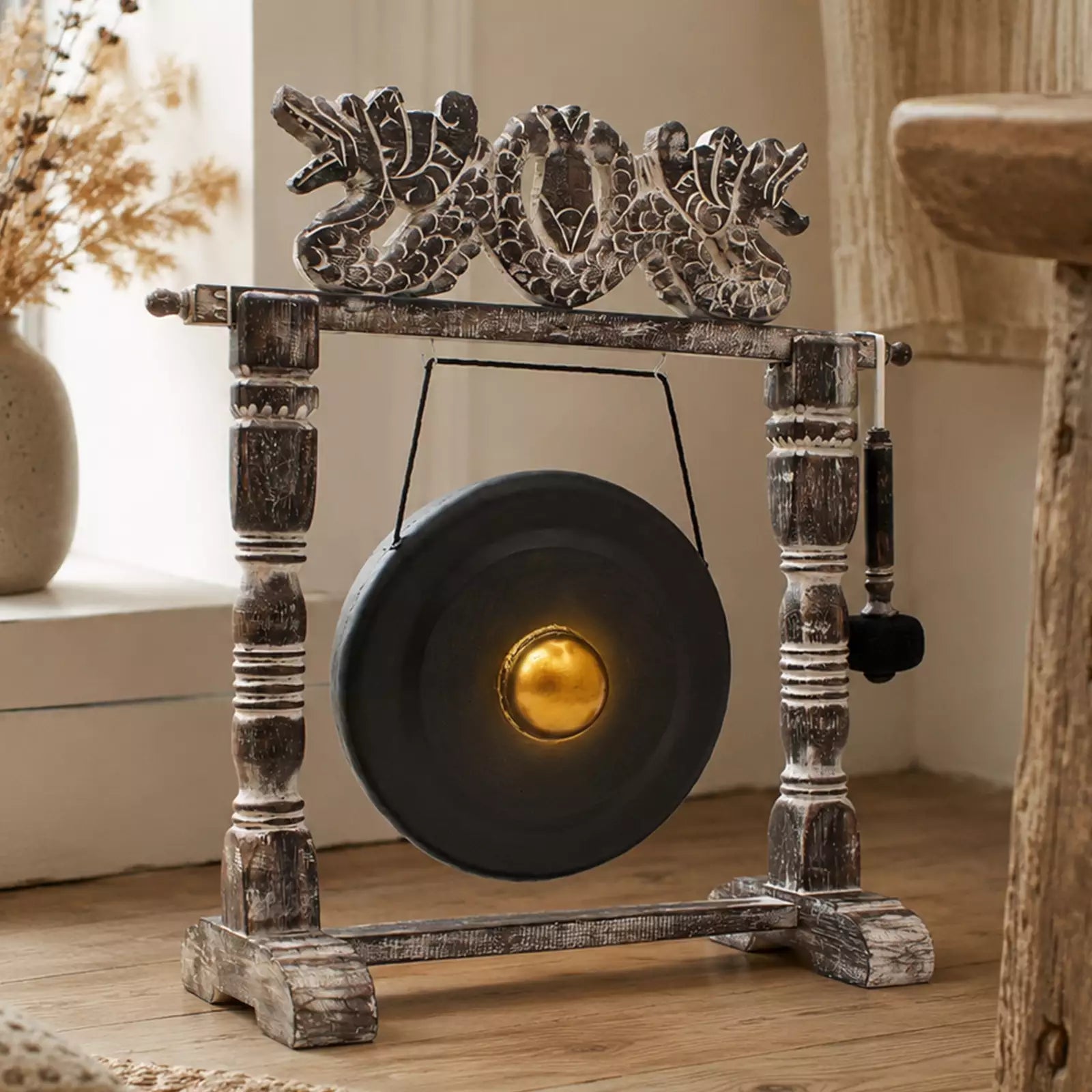 A rustic decorative gong set with a dark round gong, weathered wood stand, carved dragon-like top piece, and a hanging mallet, shown in a warm indoor setting.
