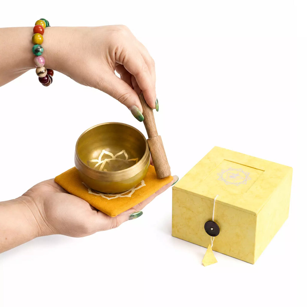A brass singing bowl set with a wooden mallet resting on a yellow cloth, shown with its yellow gift box on a white background. A person's hands are holding the bowl and mallet.