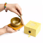 A brass singing bowl set with a wooden mallet resting on a yellow cloth, shown with its yellow gift box on a white background. A person's hands are holding the bowl and mallet.