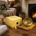 A yellow singing bowl set with a wooden mallet and matching box, styled on a wooden table in a cozy living room with a fireplace in the background.